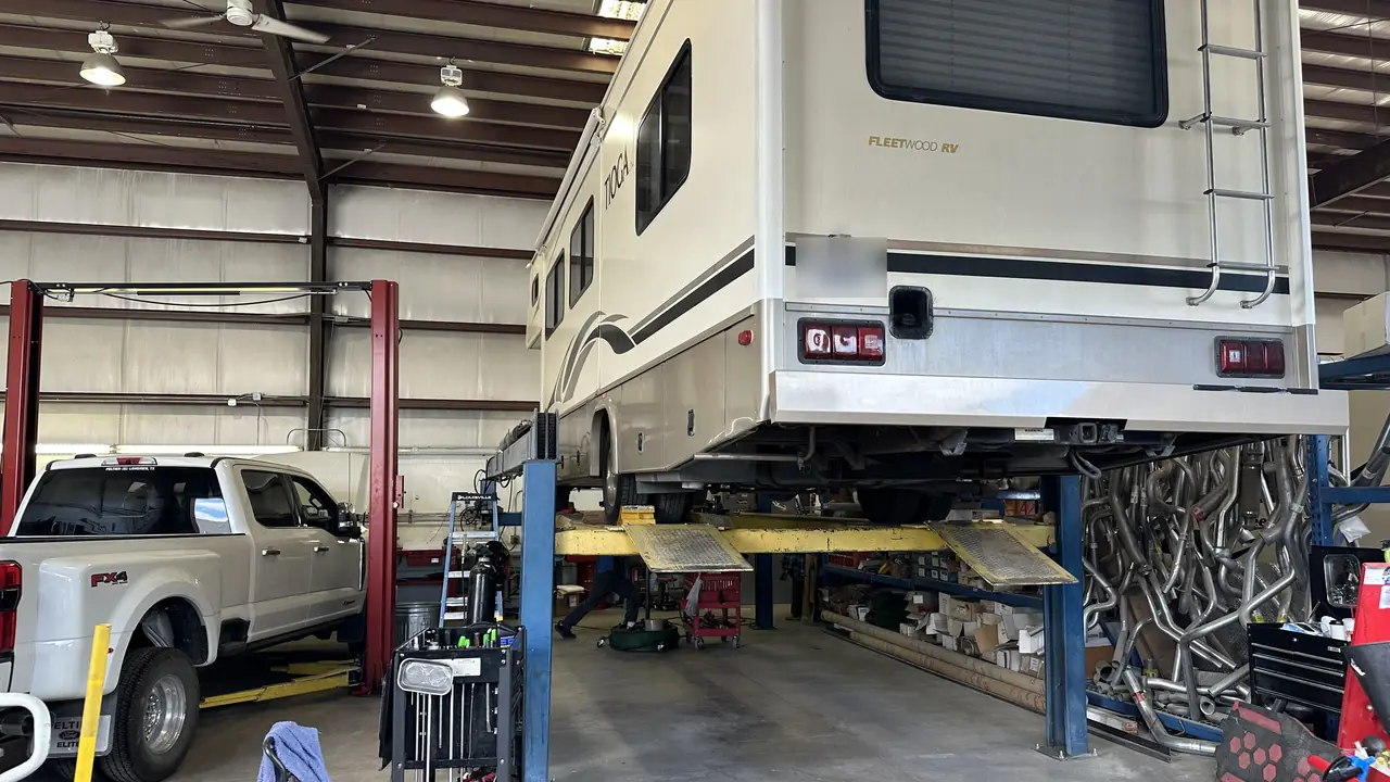 A Class C motorhome raised off the shop floor indoors, with a technician working underneath it